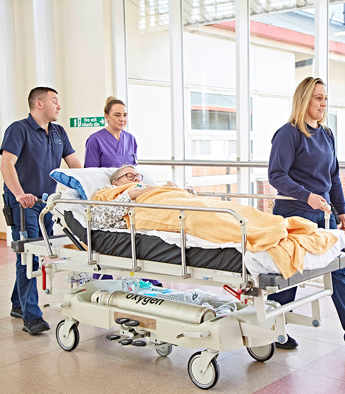Female nurse pushing patient in bed through hospital corridoor with colleague walking beside 