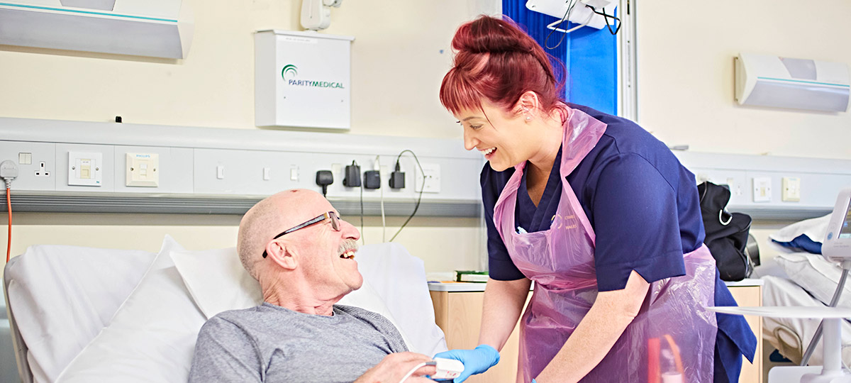 Female nurse smiling at older male patient in hospital bed 