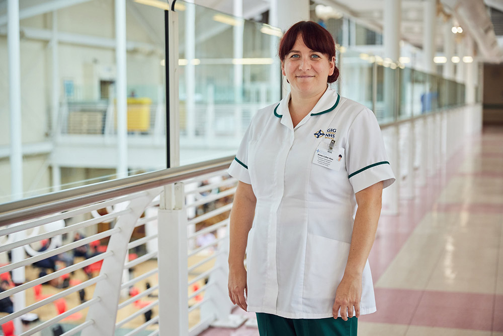 Healthcare support worker smiling at the cameria, with hospital corridor in the background 