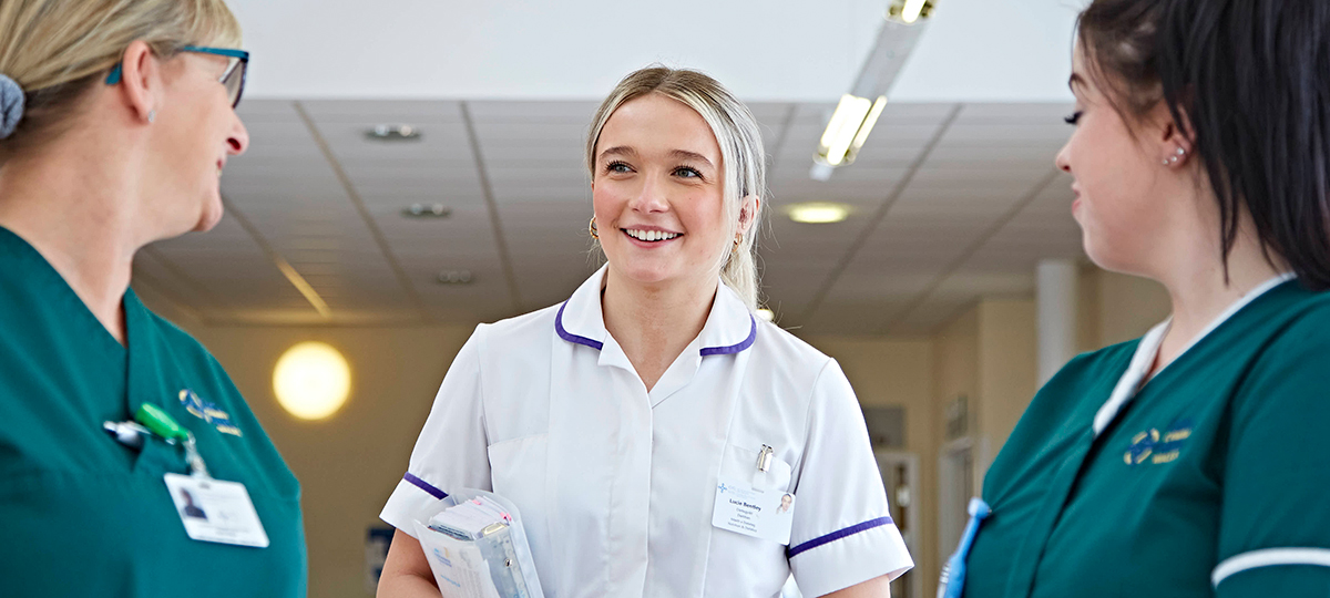 Woman in the centre of the frame, in a healthcare support worker uniform, speaking to two nurse colleagues either side of her