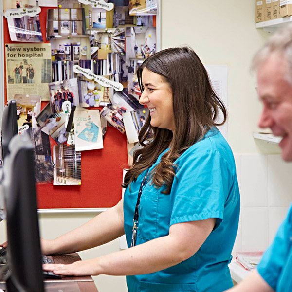 Woman stood behind desk and typing while smiling forwards 