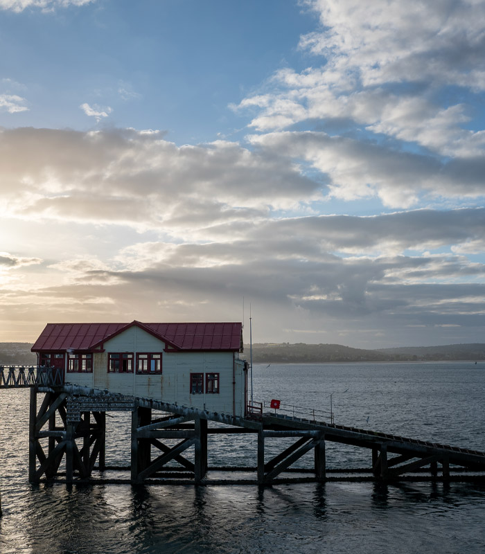 The sun setting in the background, in the foreground we see a boat house with structure in the sea 